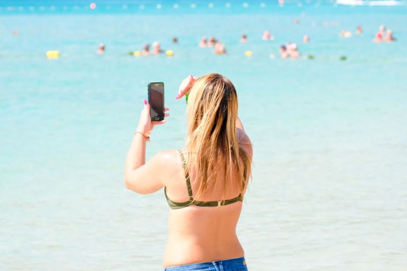 Rear view of woman photographing while shielding eyes on beach