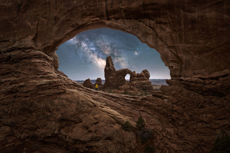 Unrecognizable traveler admiring breathtaking view of turret arch located near rough canyon against glowing milky way at night sky in arches national park in utah, usa