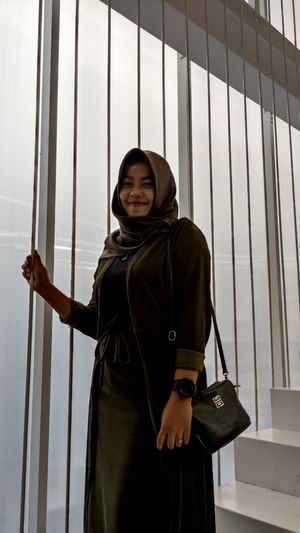 Portrait of smiling young woman wearing hijab standing by window
