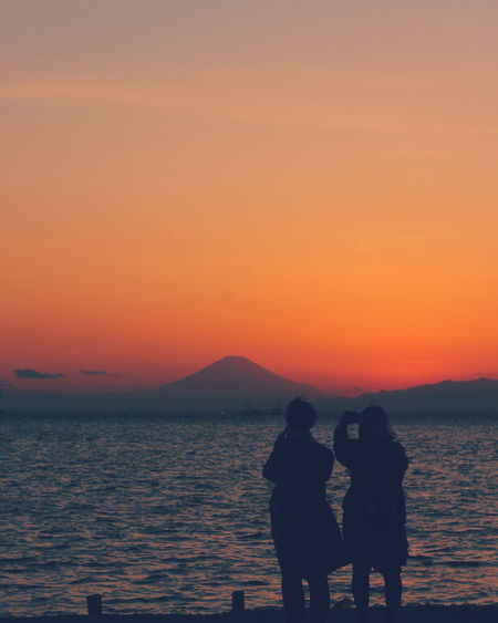 Rear view of silhouette couple standing at beach against orange sky