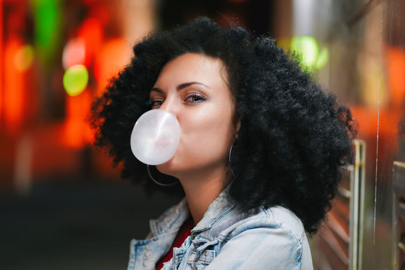 Close-up portrait of smiling young woman blowing bubble gum at night