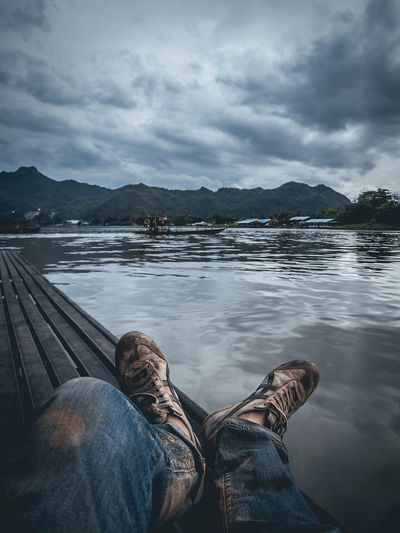 Low section of man relaxing on pier over lake against cloudy sky