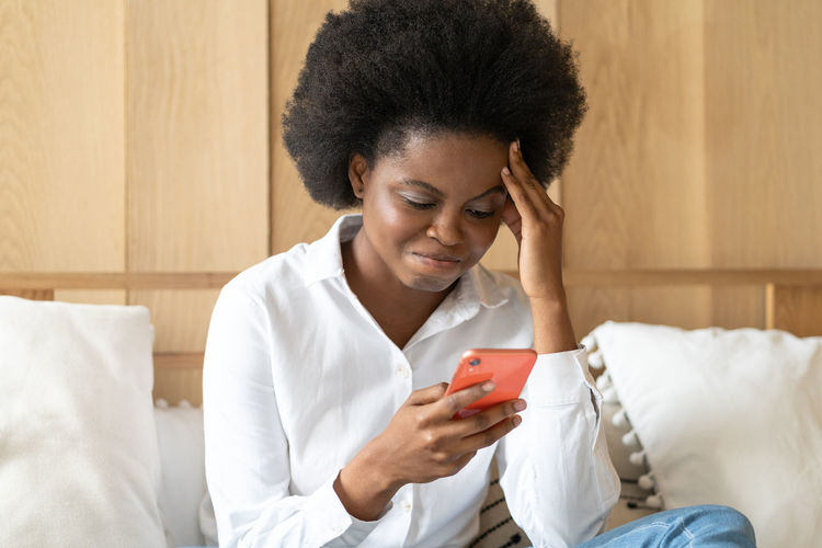 Frustrated woman using smart phone while sitting on bed at home