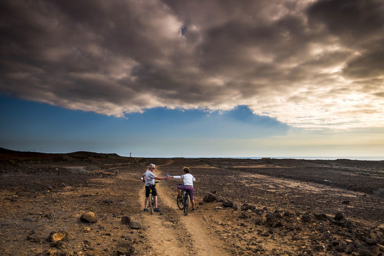 Rear view of senior couple cycling on land against cloudy sky