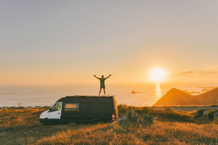 Young man on sprinter van with arms wide open in front of sunset on cliff in baja, mexico.