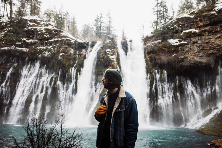 Man standing on picturesque scenery of powerful waterfall with pool flowing among snowy forest in mountainous terrain in winter day in usa looking away