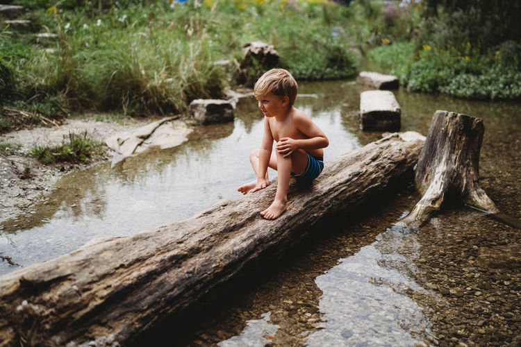 Young boy sitting on log in water in summer looking thoughtful