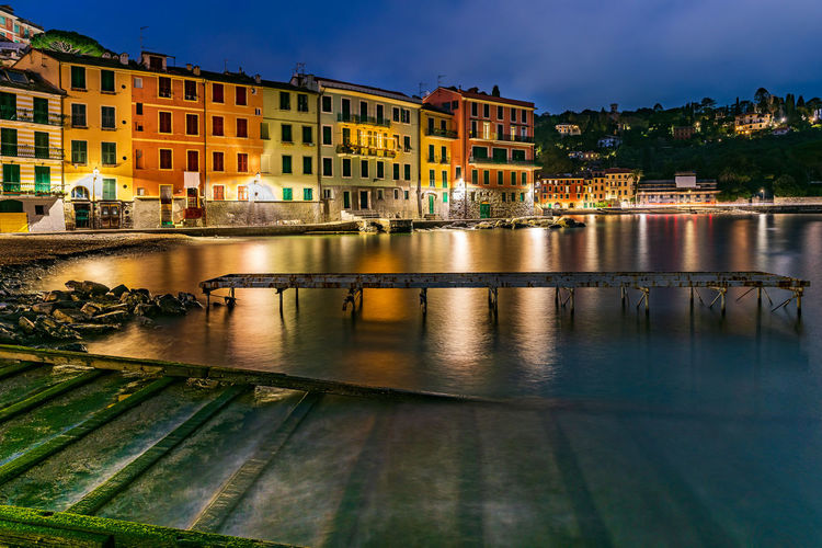 Rusty pier on lake against buildings in illuminated city at night