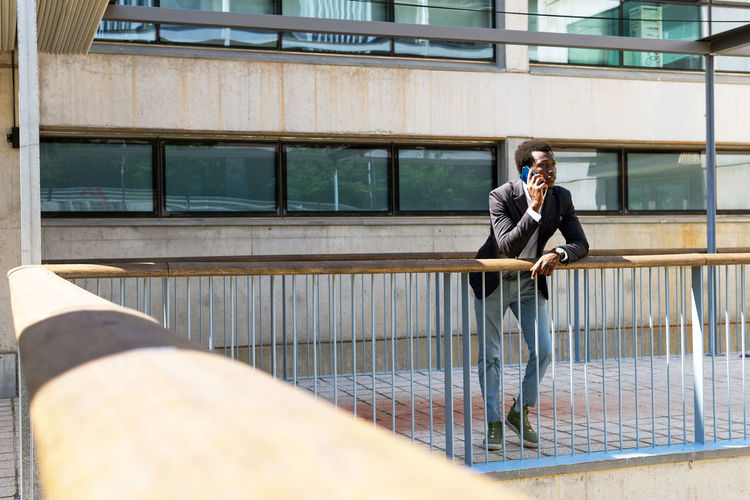 Busy african american male entrepreneur standing near office building and talking about project via smartphone while leaning on metal railing and looking away