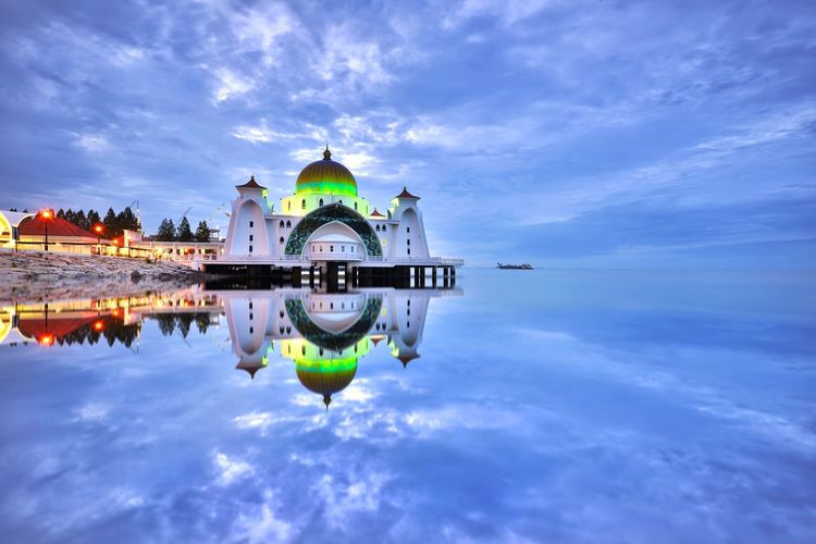 Illuminated mosque reflecting on calm sea against cloudy sky during sunset