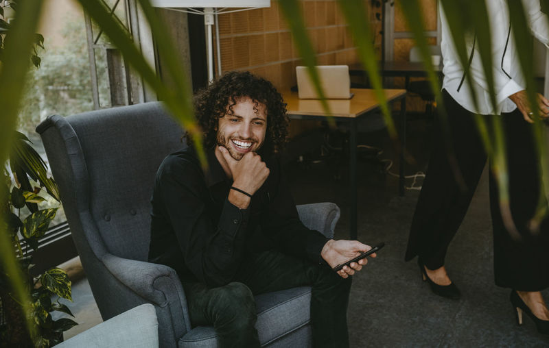 Smiling male entrepreneur holding smart phone sitting on chair in office