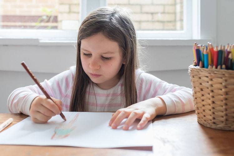 Girl drawing on paper while sitting on table at home
