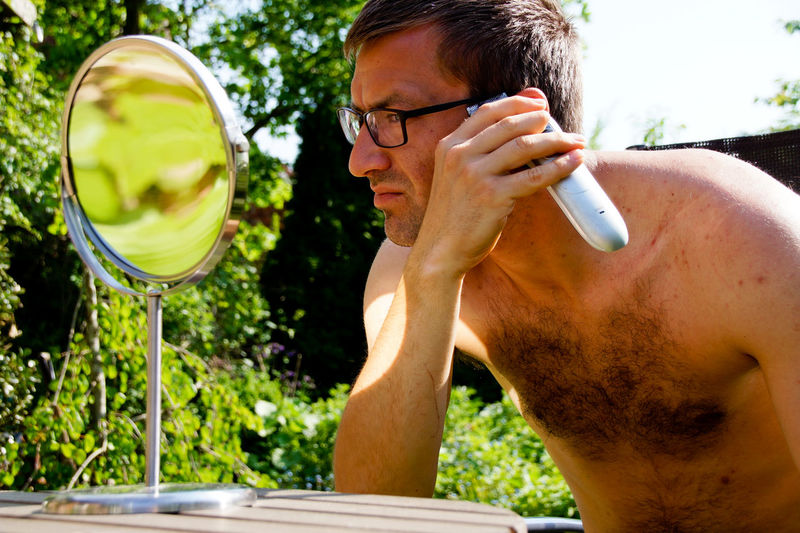 Shirtless of mid adult man shaving while looking at mirror in garden