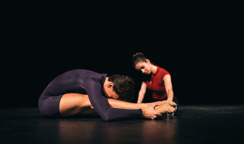 Full body male and female ballet dancers stretching on dark stage during rehearsal in theater