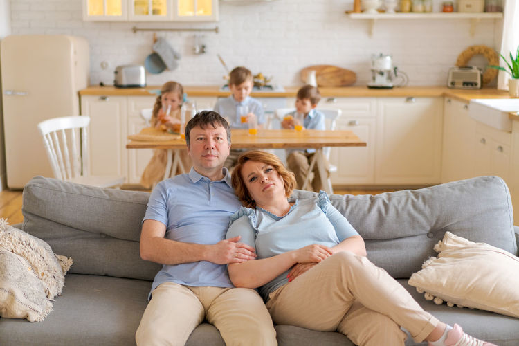 Tired parents suffer from headaches, sit on couch, while their daughter and two