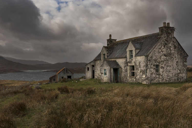 Abandoned house on grassy field by river against cloudy sky
