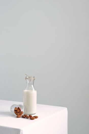 Almond milk in transparent glass bottle and pile of roast almonds on white table against white wall