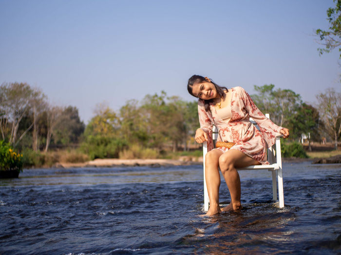 Woman smiling while sitting by water against clear sky