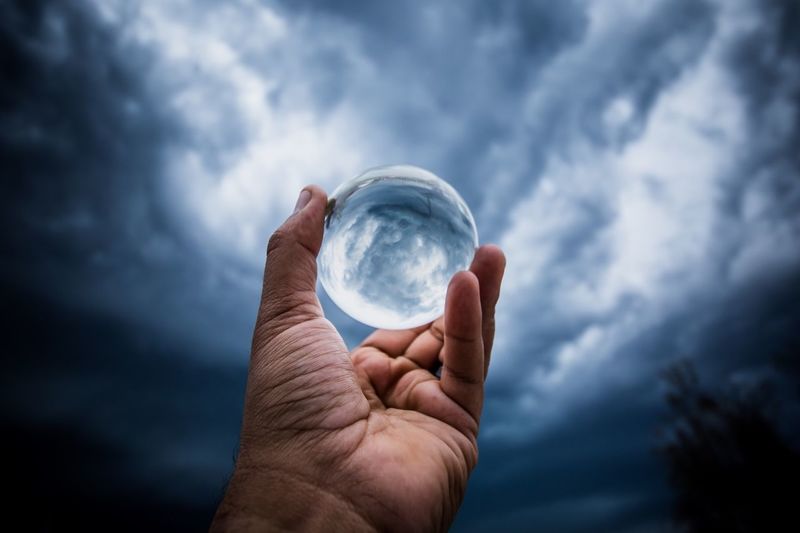 Close-up of hand holding crystal ball against cloudy sky