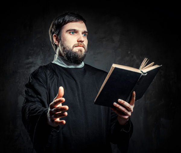 Priest holding bible while standing against black background