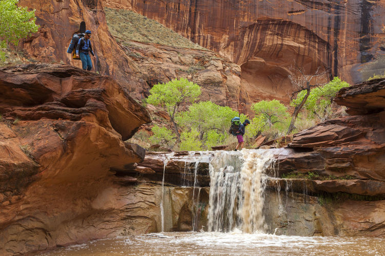 People hike across waterfall in sandstone canyon, coyote gulch, utah