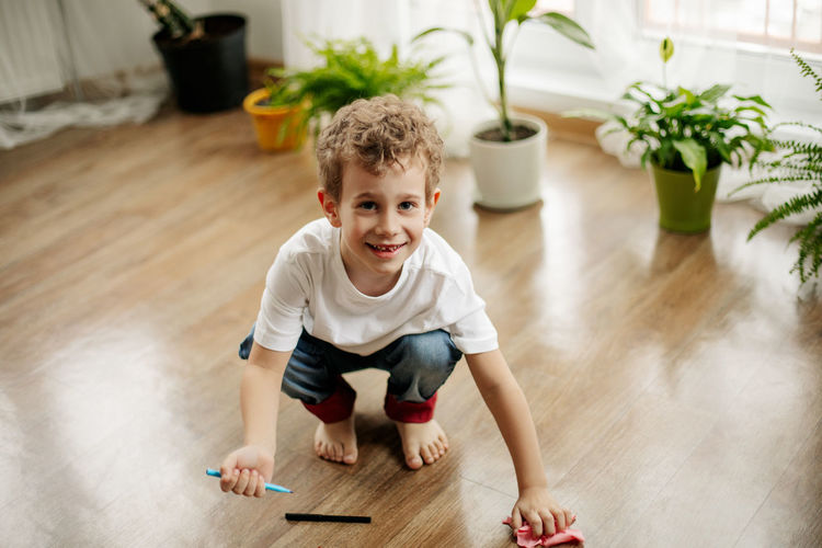 The boy is squatting on the floor in the room, holding a marker in his hand, wiping the floor