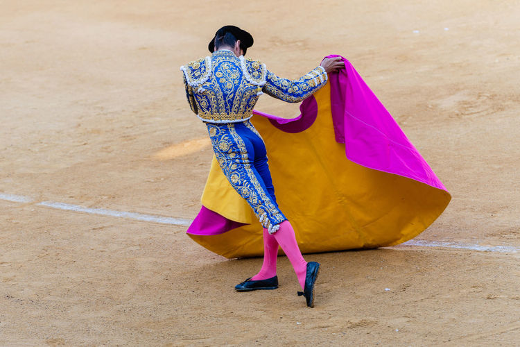 Back view of anonymous toreador in bright traditional costume and with cloak performing on sandy bullring during corrida festival