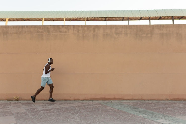 Side view full body of energetic african american sportsman in headphones jogging along pavement during fitness training