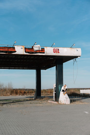 Young woman sitting at fuel pump against sky