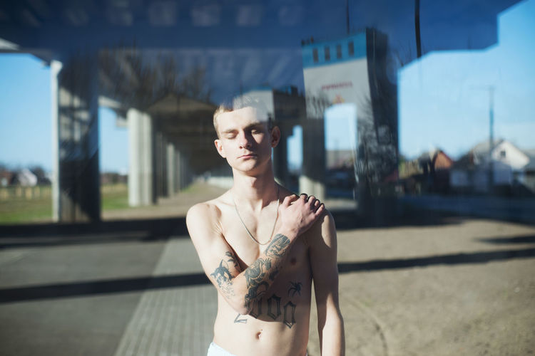 Shirtless young man standing outdoors seen through glass