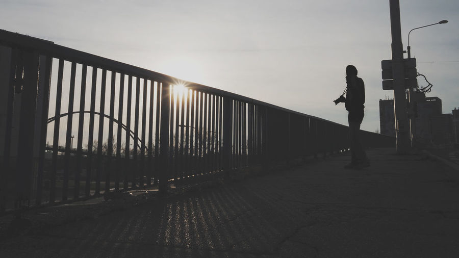 Man walking on footbridge against sky during sunset