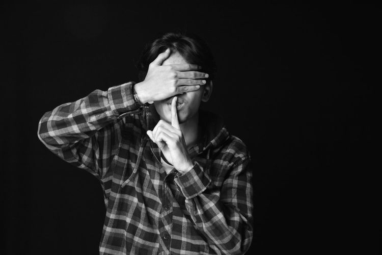 Young man with finger on lips covering eyes against black background