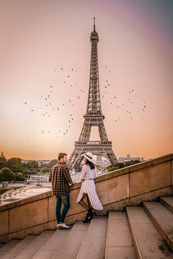 Full length of couple standing against eiffel tower on steps in city during sunset