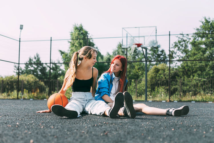 Two girls in sports clothes and with a basketball are chatting, sitting on the playground.