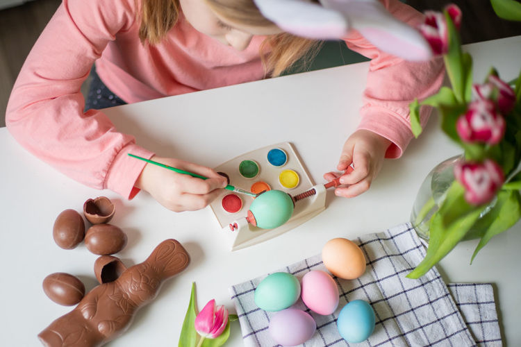 Top view of a little girl with bunny ears holding paintbrush and decorating easter eggs watercolors.