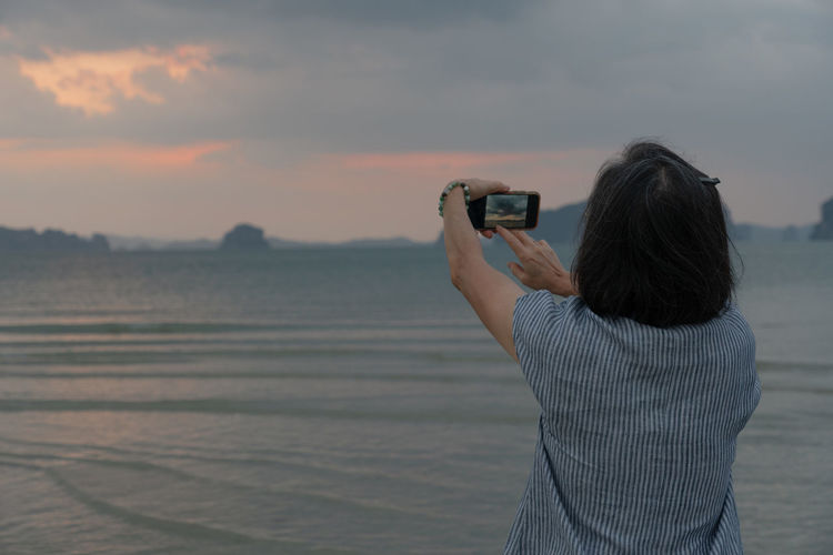 Senior woman using smartphone to take photo of ocean at sunrise during summer vacation