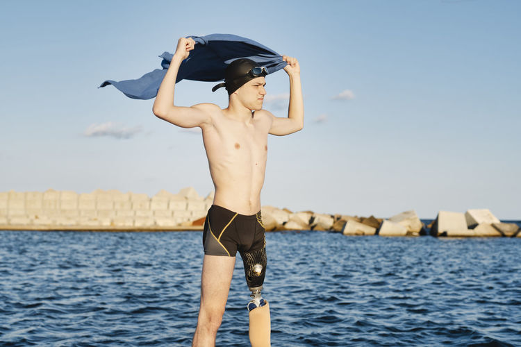 Young man standing with towel against sea on sunny day