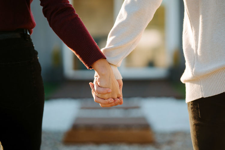 Crop anonymous couple tenderly holding hands while standing on blurred background of house on sunny day