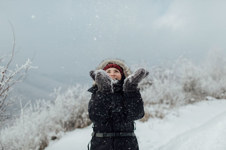 Cheerful woman in warm clothing throwing snow while standing on mountain