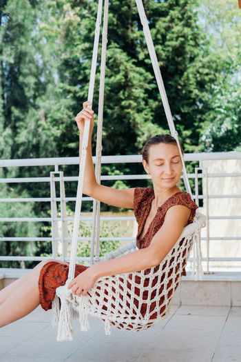 Young boho slyle charming woman sitting with closing eyes in hammock on balcony her house.