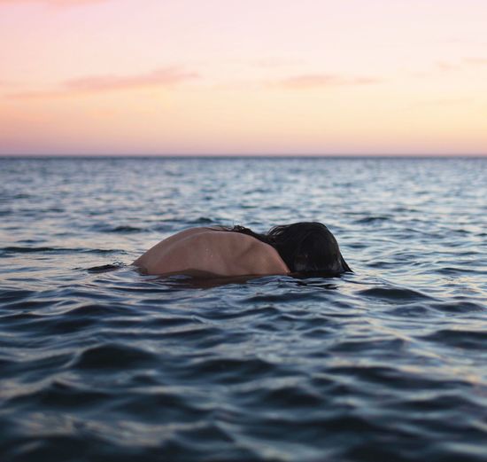 Shirtless person swimming in sea against sky during sunset