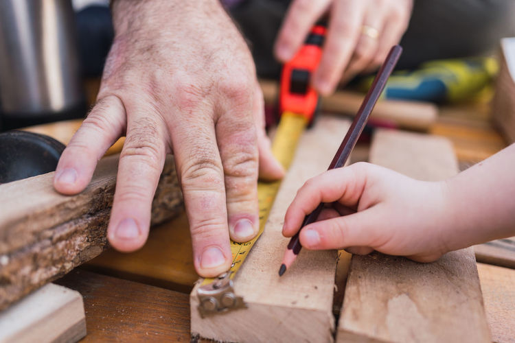 Crop unrecognizable dad measuring wooden piece with tape against child with pencil sitting on boardwalk