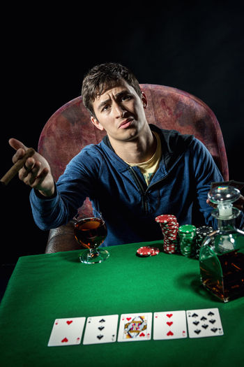 Portrait of sad man sitting at poker table with whiskey and cigar against black background