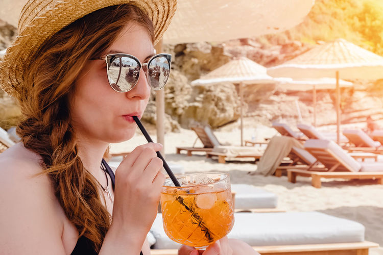 Woman in straw hat and sunglasses drinking aperol spritz cocktail on the beach. 