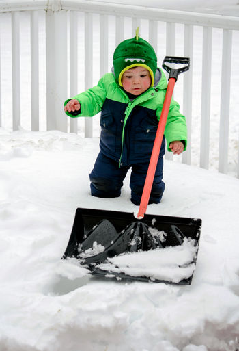 A toddler boy stands in the snow looking at a very big snow shovel. 