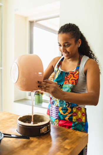 Happy ethnic african american female in apron pouring liquid chocolate batter into baking form with baking paper