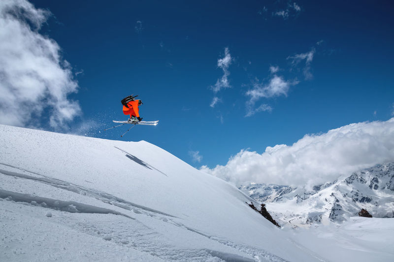 A freerider skier in an orange suit with a backpack froze in a jump flight over high snow-capped 