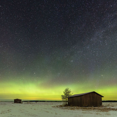 Wooden houses on snowcapped field against aurora borealis in sky