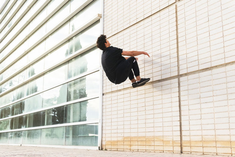 Side view of full length man in black outfit performing acrobatic parkour on wall of building