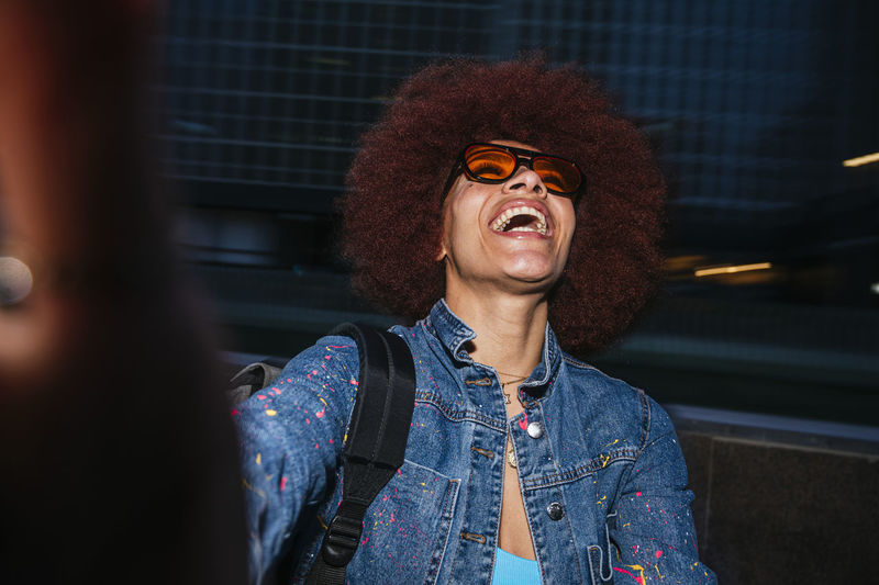 Joyful female with afro hairstyle wearing trendy denim outfit and sunglasses taking self portrait on dark street in evening time